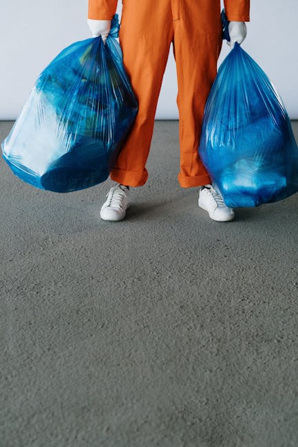 A person wearing orange work overalls and white sneakers is standing on a paved surface, holding two large blue plastic rubbish bags filled with waste. The bags are semi-transparent, revealing various