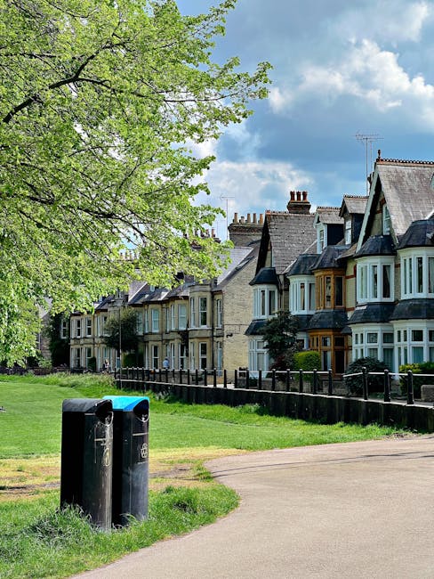A suburban street scene featuring a row of Victorian-style terraced houses with bay windows, painted in muted tones of beige, cream, and light blue, situated behind a well-maintained black metal fence